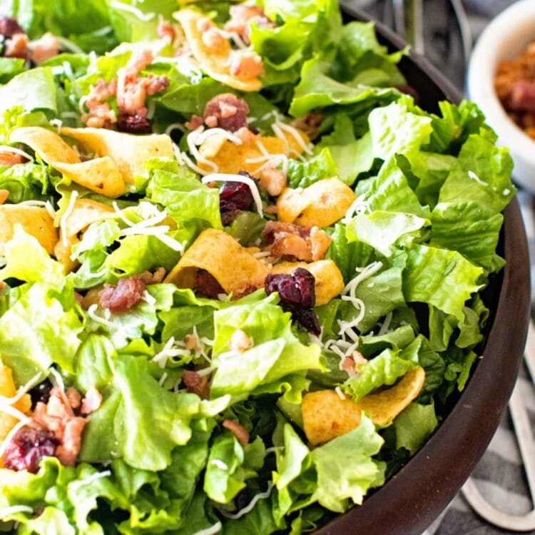 Lettuce Frito Salad in a wood bowl next to a grey and white striped towel with a metal tongs