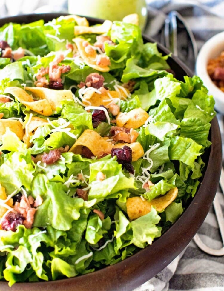 Lettuce Frito Salad in a wood bowl next to a grey and white striped towel with a metal tongs