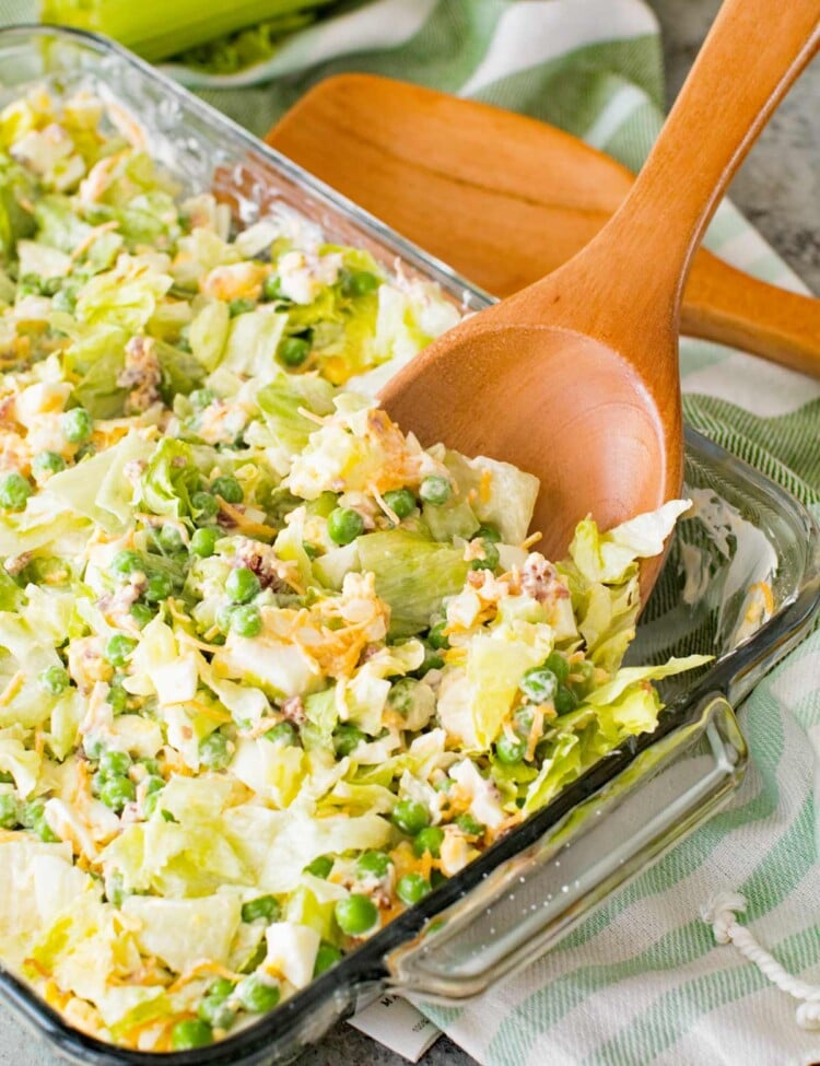 Traditional Seven Layer Salad in a clear glass baking dish with a wood spoon in it sitting on a white and green striped kitchen towel