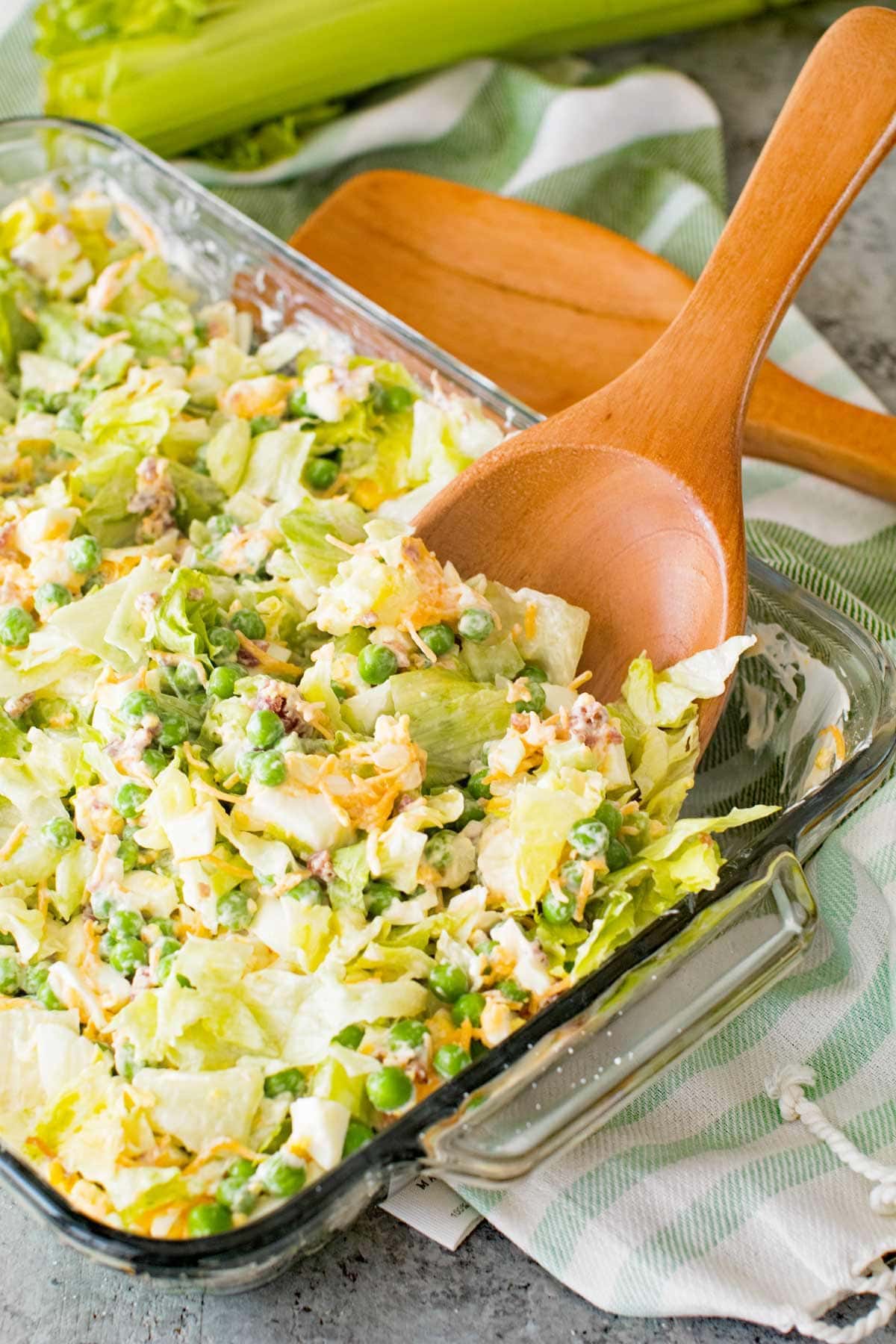 Traditional Seven Layer Salad in a clear glass baking dish with a wood spoon in it sitting on a white and green striped kitchen towel