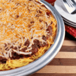 A metal pan of spaghetti pie on a wood board along with a stack of white plates and a spatula