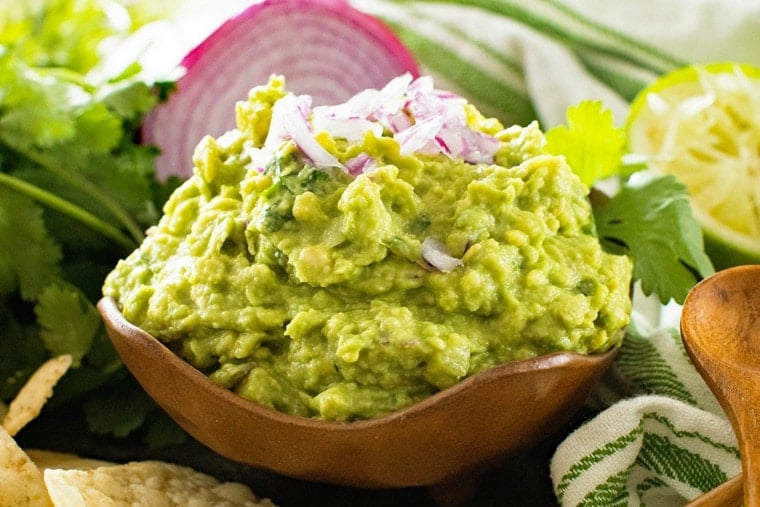 Guacamole in brown bowl with red onion cilantro lime and chips in background