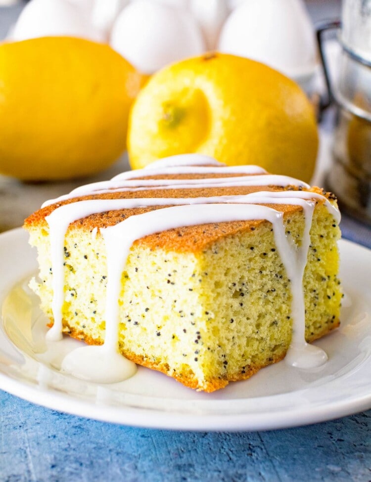 Poppy seed cake piece on a white plate with lemons and eggs in the background