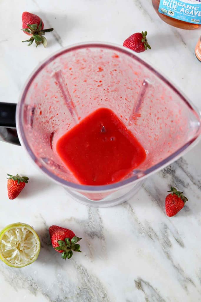 A blender, full of frozen Strawberry Virgin Margarita, sits on a marble countertop, surrounded by strawberries and juiced limes.