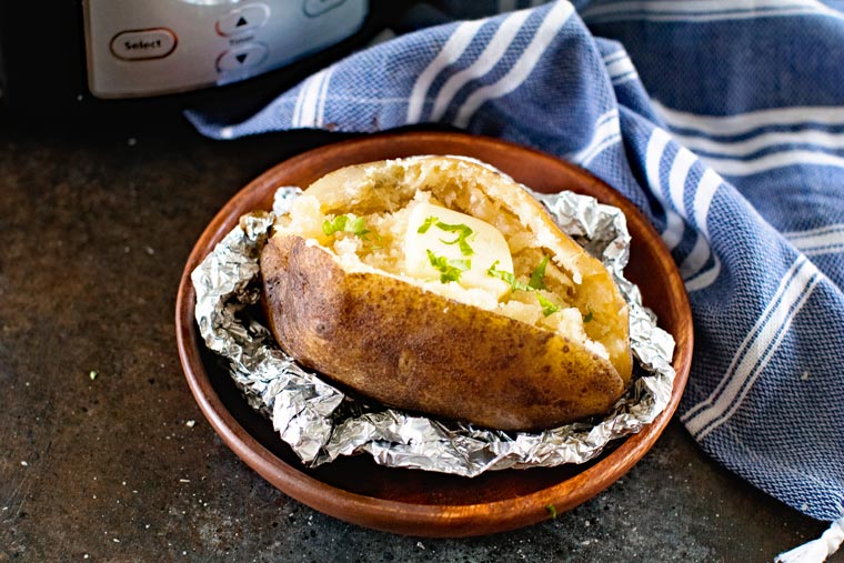 Wooden circular plate with baked potato cut open on a piece of foil with butter on top.