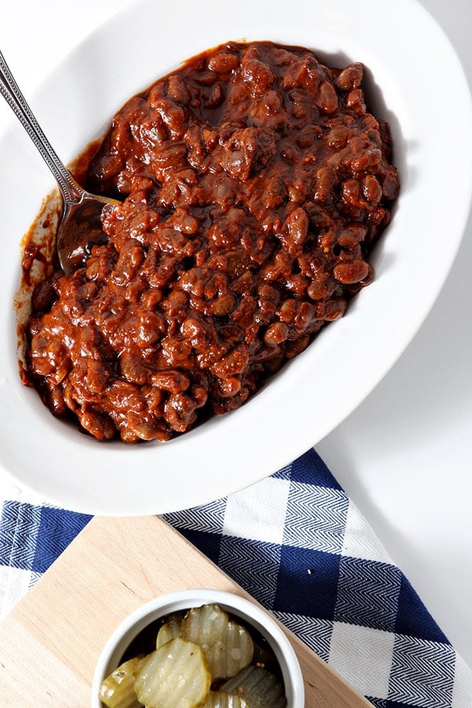 Overhead image of Instant Pot Baked Beans, shown in a white bowl, shown with a blue buffalo plaid napkin and a platter of barbecue sides