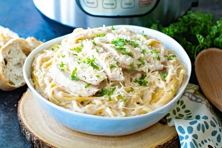 Bowl of Instant Pot Chicken Alfredo on piece of wood with napkin, wooden spoon and pressure cooker in background.