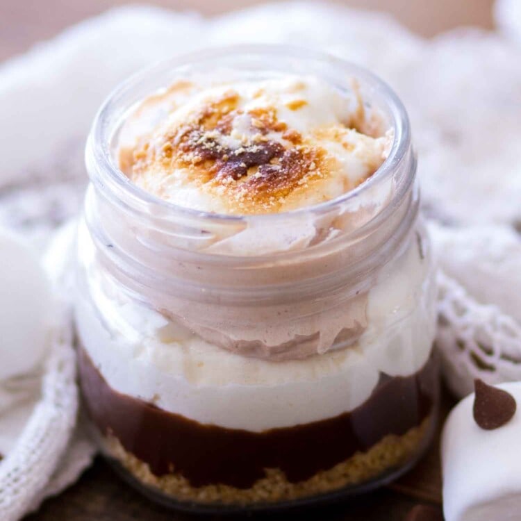 Mason jar of no bake s'mores trifle on a whood table with chocolate chips and marshmallows