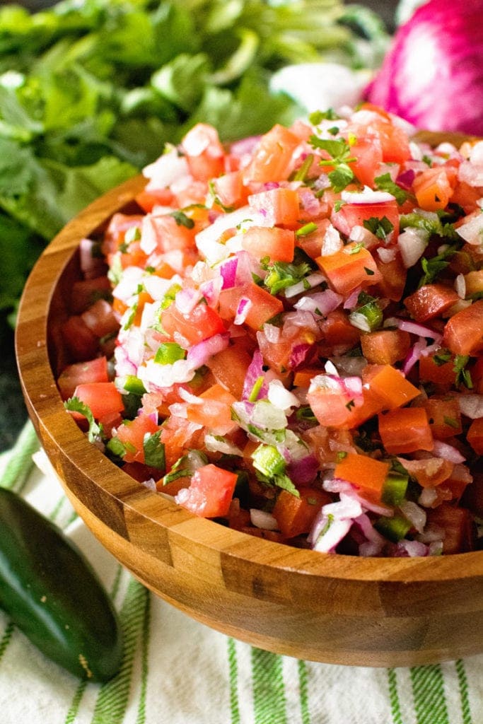 Wooden bowl filled with pico de gallo on a green and white striped napkin. A bunch of cilantro, red onion and jalapeno in background.