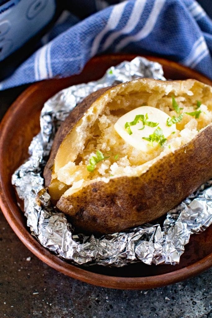 Close up image of a sliced open baked potato topped with butter on a scrunched piece of aluminum foil. A blue and white striped cloth napkin in background.