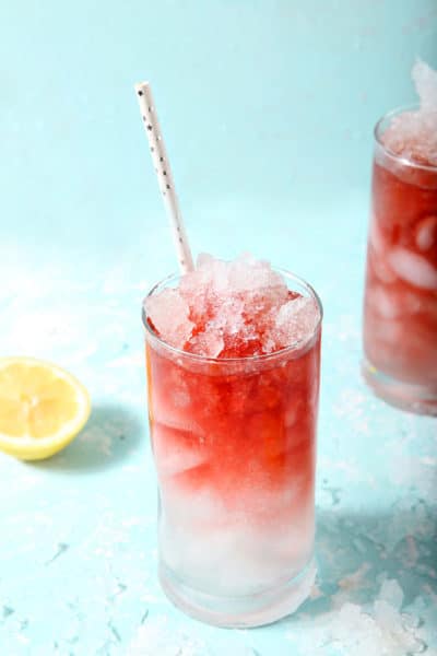 Two glasses of Arnold Palmer Iced Tea are surrounded by ice and lemon slices on a teal background