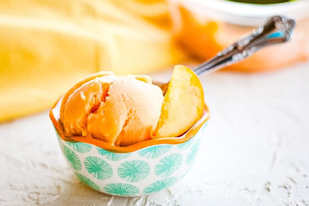 White background with bowl of peach sorbet with spoon on it and orange napkin in background