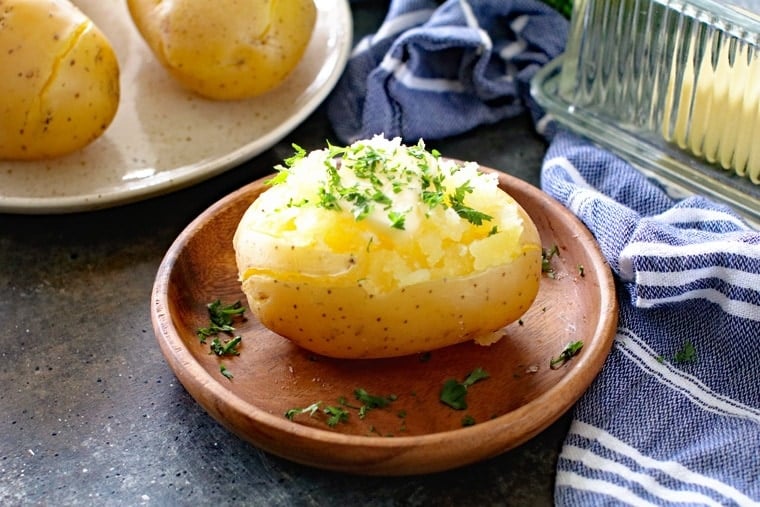 Baked Potato on brown plate with more in the background and a glass butter dish behind it