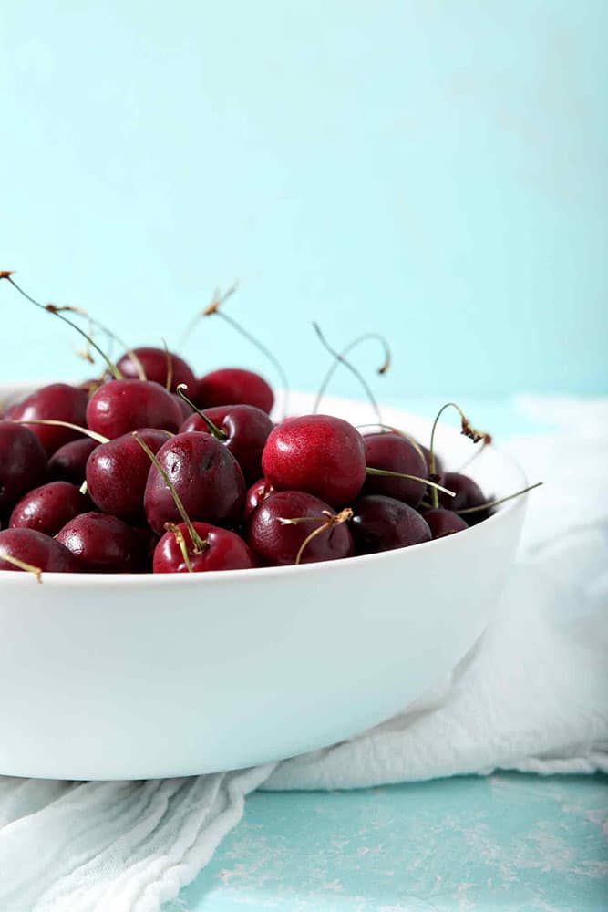 Cherries sit in a white bowl on a turquoise background