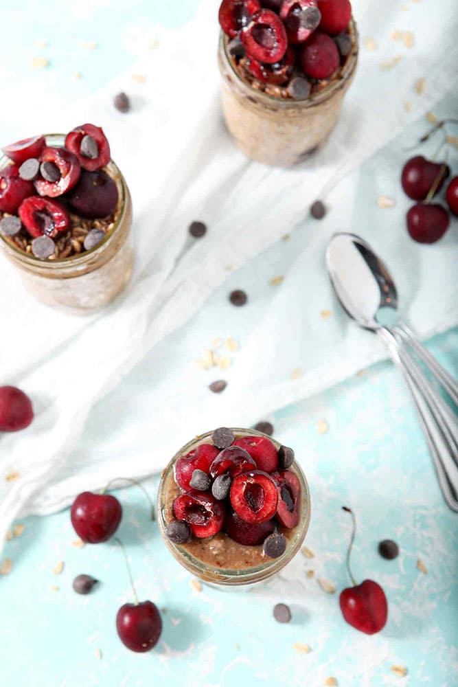 Overhead image of Cherry Overnight Oats Recipe, surrounded by more fresh cherries and chocolate chips, as they sit on a white towel on a turquoise background