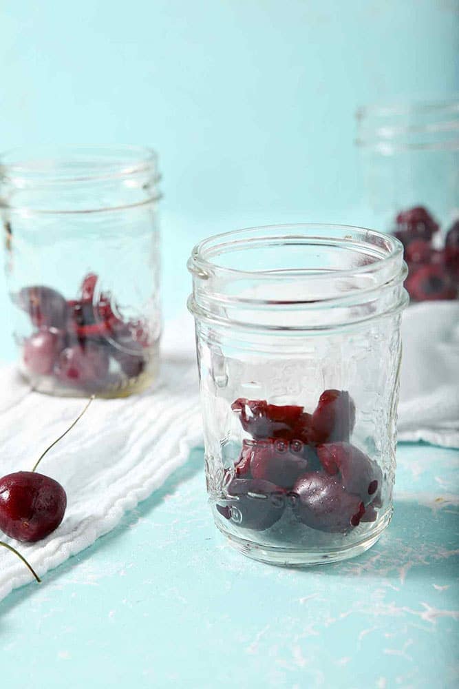 Halved, pitted cherries are shown in the base of mason jars as cherry overnight oats are assembled