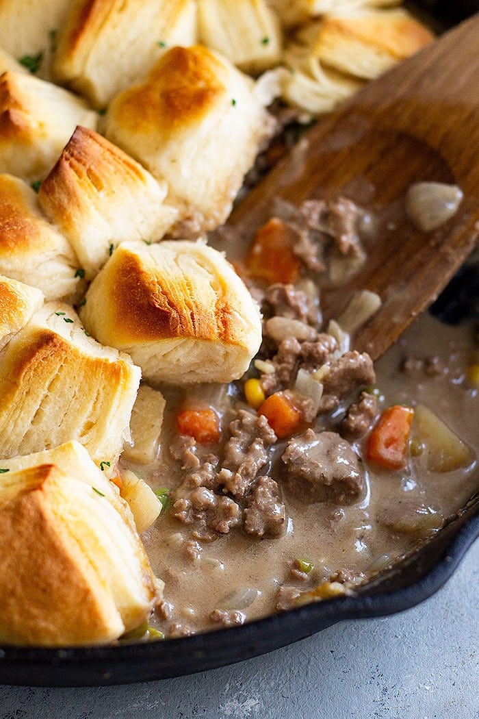 Ground beef pot pie in a skillet with biscuits. A wooden spoon is mixing the Beef Pot Pie