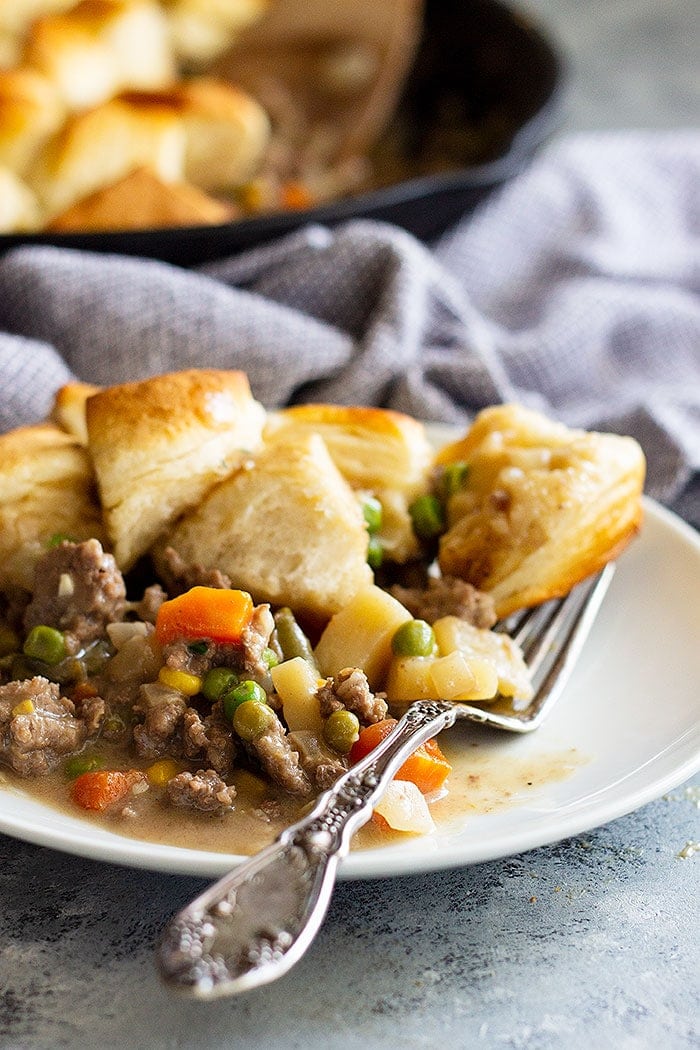 Beef Pot Pie Recipe on a white plate topped with biscuits. A cast iron skillet is in the background filled with more ground beef pot pie.