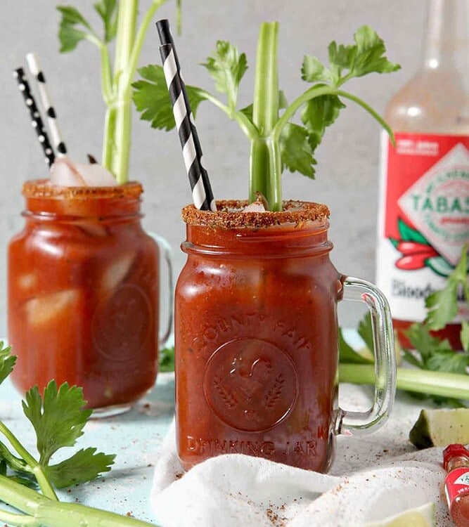 Two BBQ Bloody Marys sit next to each other with Bloody Mary Mix in the background, surrounded by limes and celery stalks