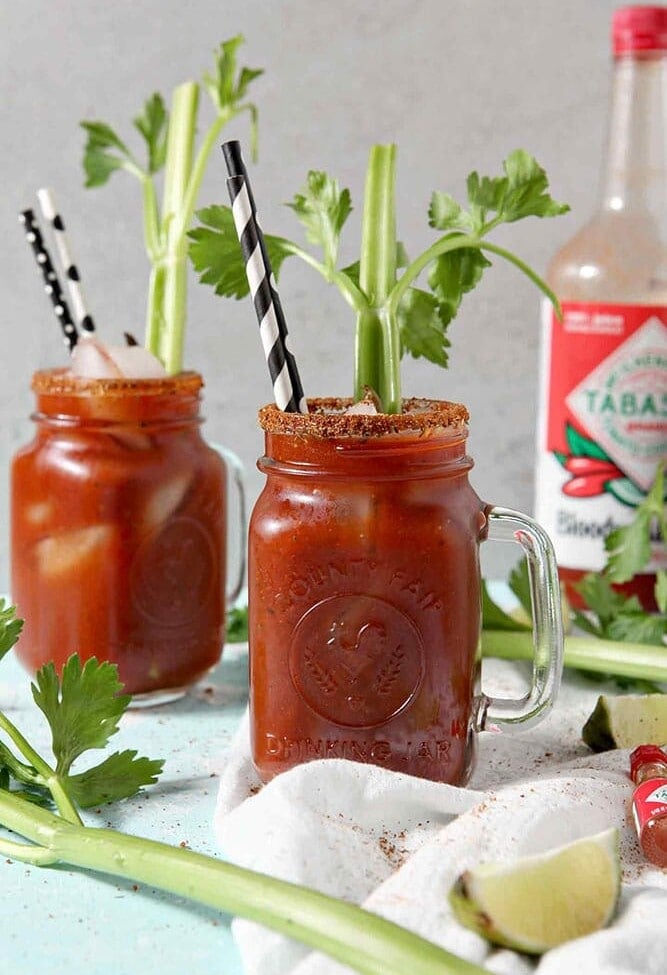 Two BBQ Bloody Marys sit next to each other with Bloody Mary Mix in the background, surrounded by limes and celery stalks