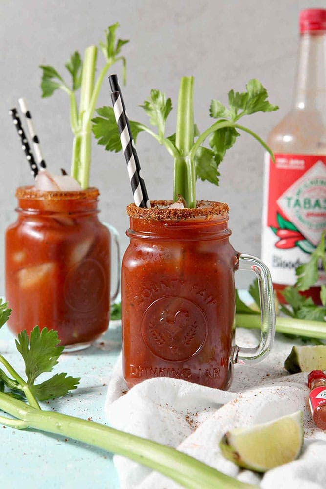 Two BBQ Bloody Marys sit next to each other with Bloody Mary Mix in the background, surrounded by limes and celery stalks