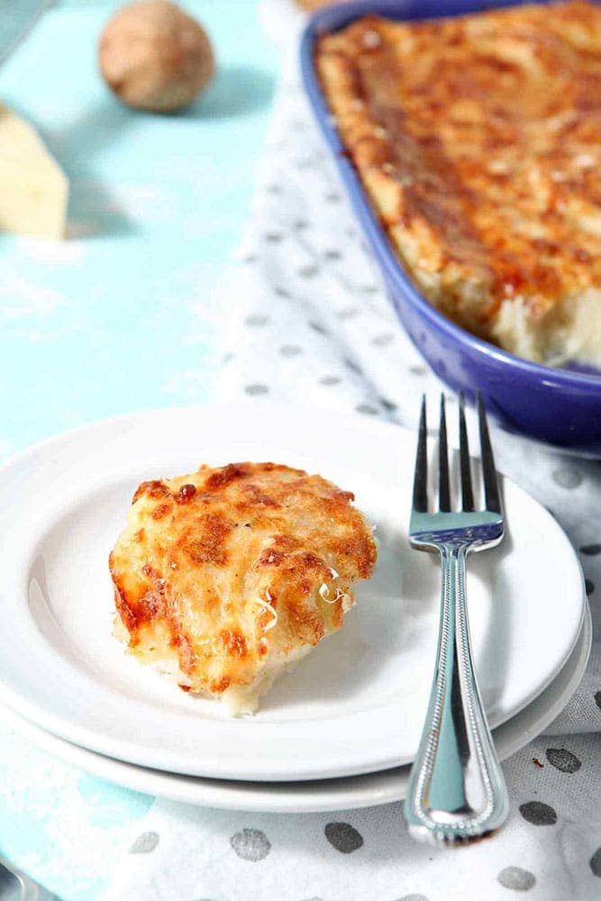 Make Ahead Mashed Potato Casserole is served on a white plate with the casserole dish in the background