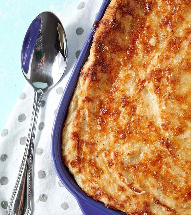 A close up of Garlic Parmesan Mashed Potato Casserole, shown in a purple baking dish with a serving spoon