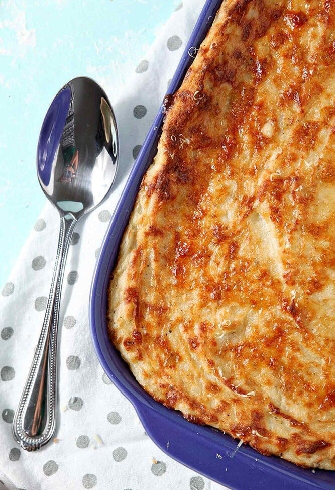 A close up of Garlic Parmesan Mashed Potato Casserole, shown in a purple baking dish with a serving spoon