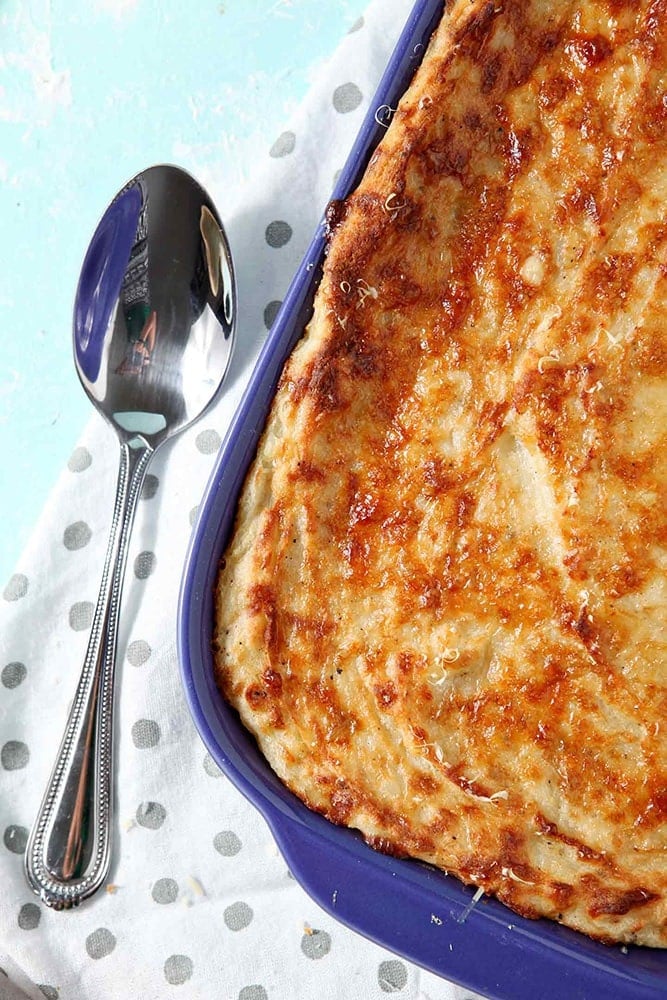 A close up of Garlic Parmesan Mashed Potato Casserole, shown in a purple baking dish with a serving spoon