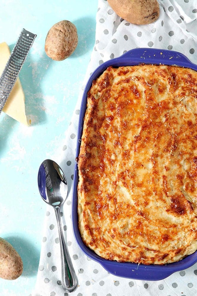 Garlic Parmesan Mashed Potato Casserole is served in its purple baking dish with a serving spoon and is surrounded by potatoes, as well as a block of parmesan cheese and a grater
