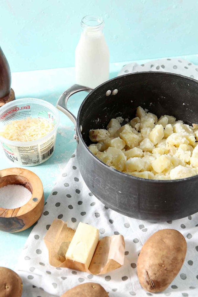 Cooked potatoes are shown in a pot, surrounded by other ingredients used in Garlic Parmesan Mashed Potato Casserole