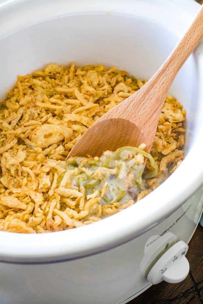 White crockpot with green bean casserole in it and a wooden spoon sitting in it.