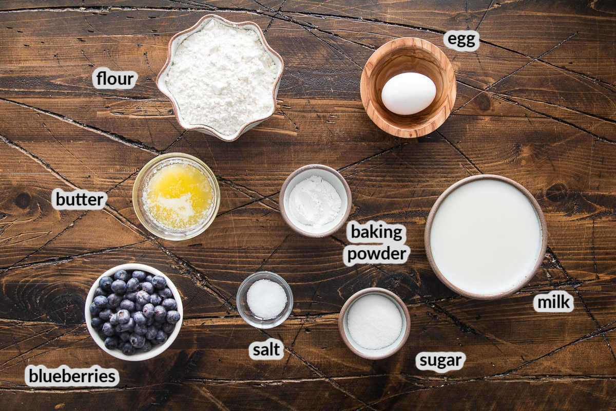 Overhead image showing ingredients to make blueberry pancakes in bowls on wood background