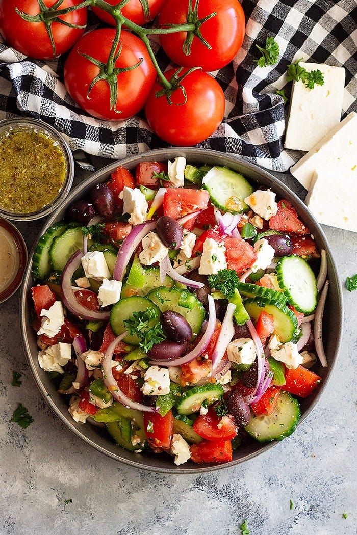 Overhead image of greek salad in bowl with fresh tomatoes, feta cheese and dressing beside it on a concrete background