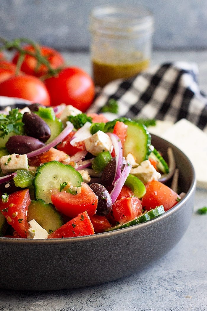 A close up photo of a gray bowl filled with Greek Salad iwth tomatoes and dressing in a mason jar in background