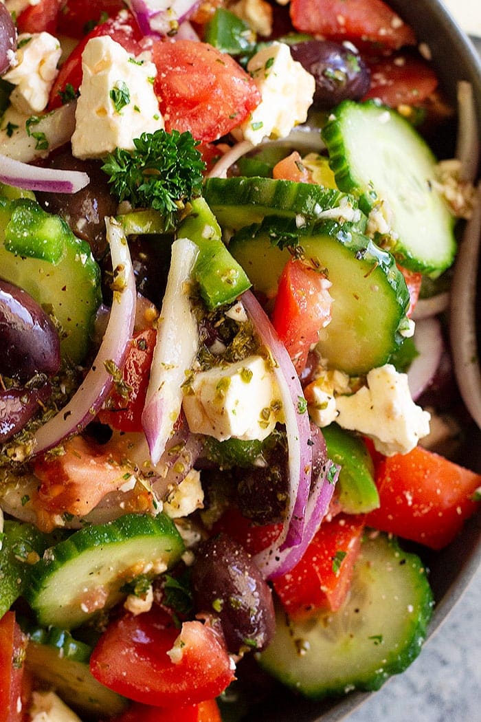 Close of photo of salad showing cucumbers, feta cheese, red onion, tomatoes and olives with dressing on it