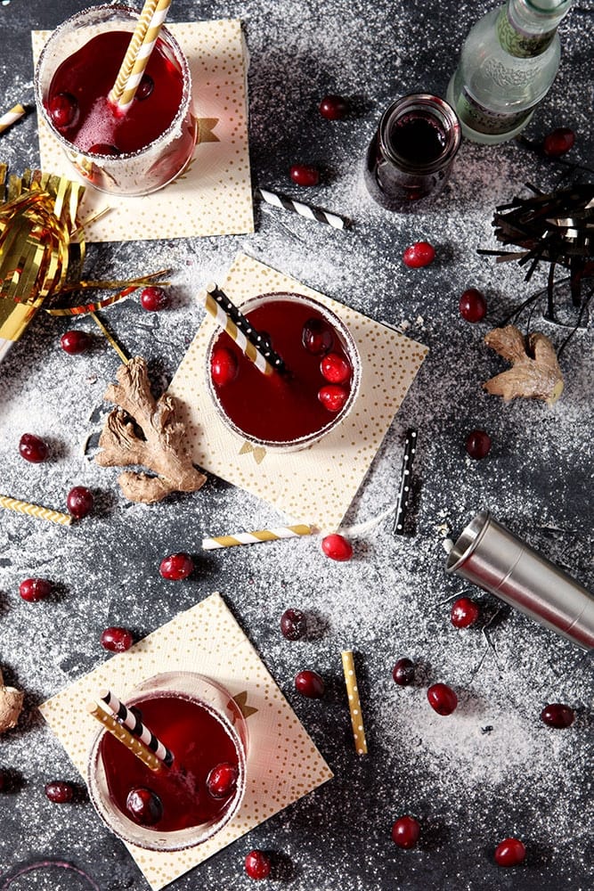Three glasses ofSparkling Ginger Cranberry Mocktails are shown from above on a black board, surrounded by cranberries, ginger and "snow"