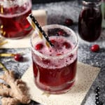 Two Sparkling Ginger Cocktails are shown on a dark background, surrounded by fresh cranberries and ginger