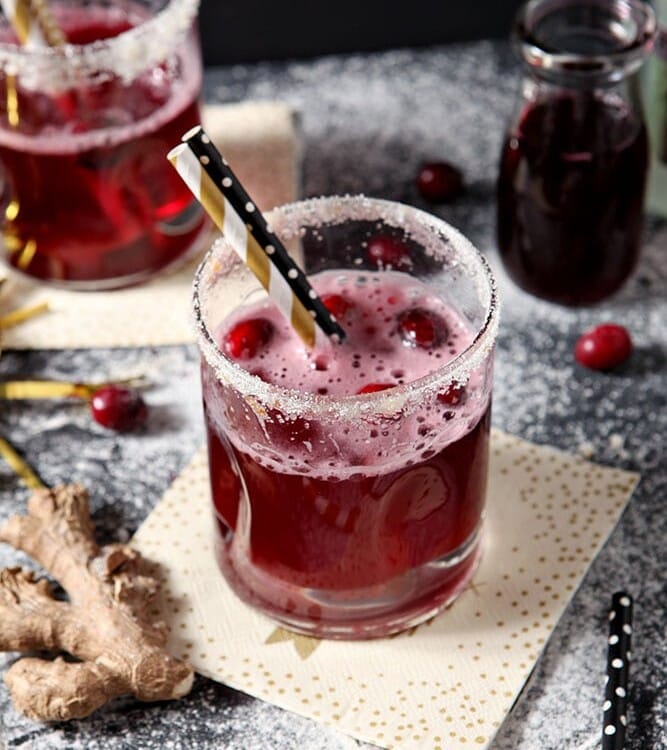 Two Sparkling Ginger Cocktails are shown on a dark background, surrounded by fresh cranberries and ginger