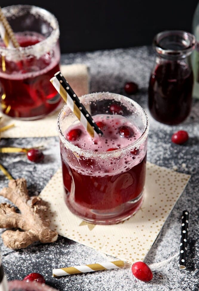 Two Sparkling Ginger Cocktails are shown on a dark background, surrounded by fresh cranberries and ginger