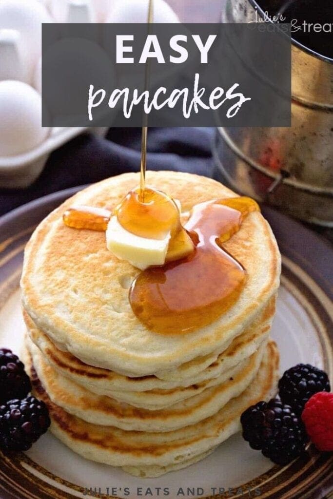 Syrup being poured over a stack of pancakes with butter on top and berries on the side