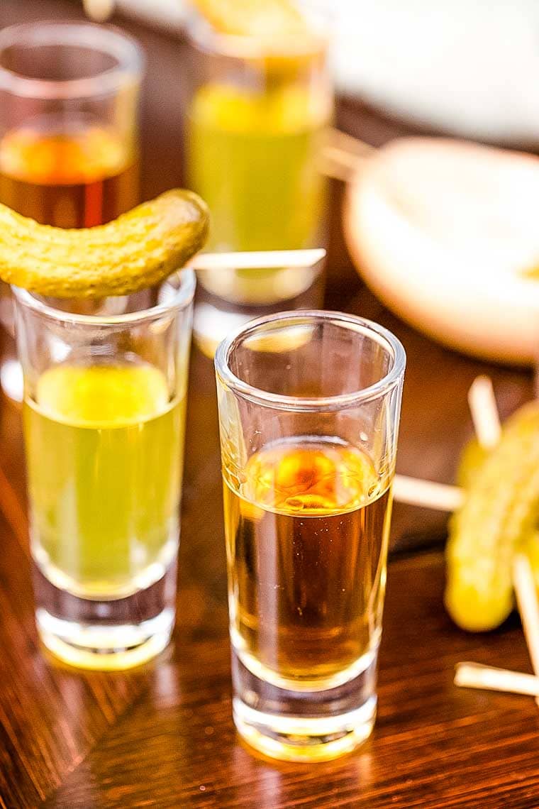 A shot glass filled with whiskey and a shot glass filled with pickle juice topped with a pickle makes a Pickleback Shot