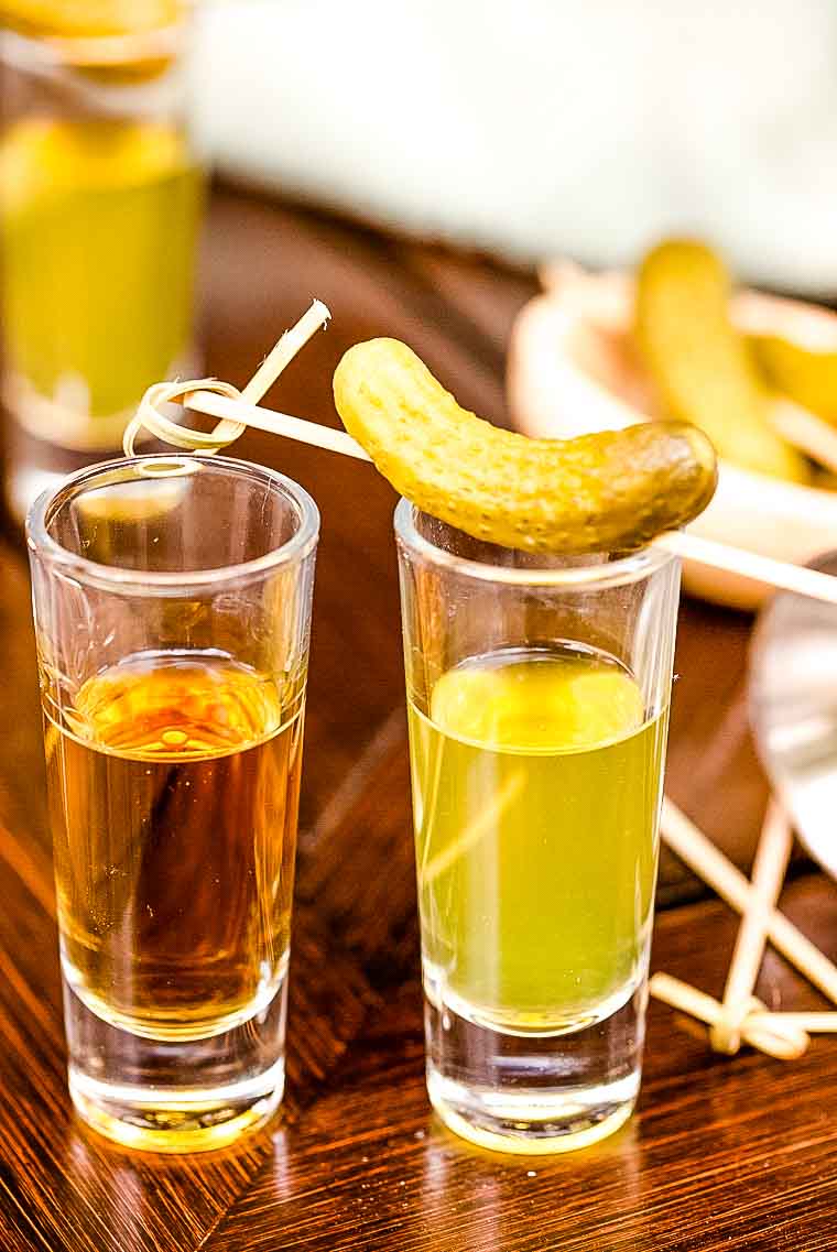 Two shot glasses with whiskey and pickle brine with a pickle garnish on top