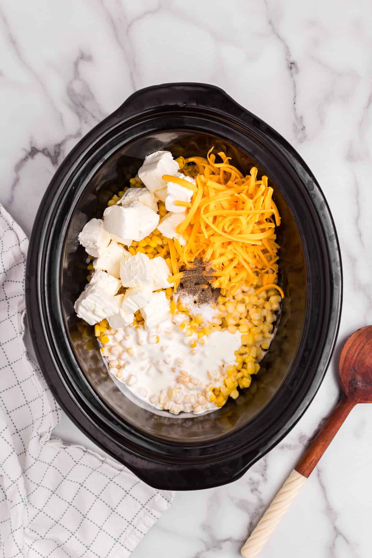 A slow cooker filled with ingredients for creamed corn including cubed cream cheese, shredded cheddar cheese, whole kernel corn, heavy cream, and seasoning before mixing.
