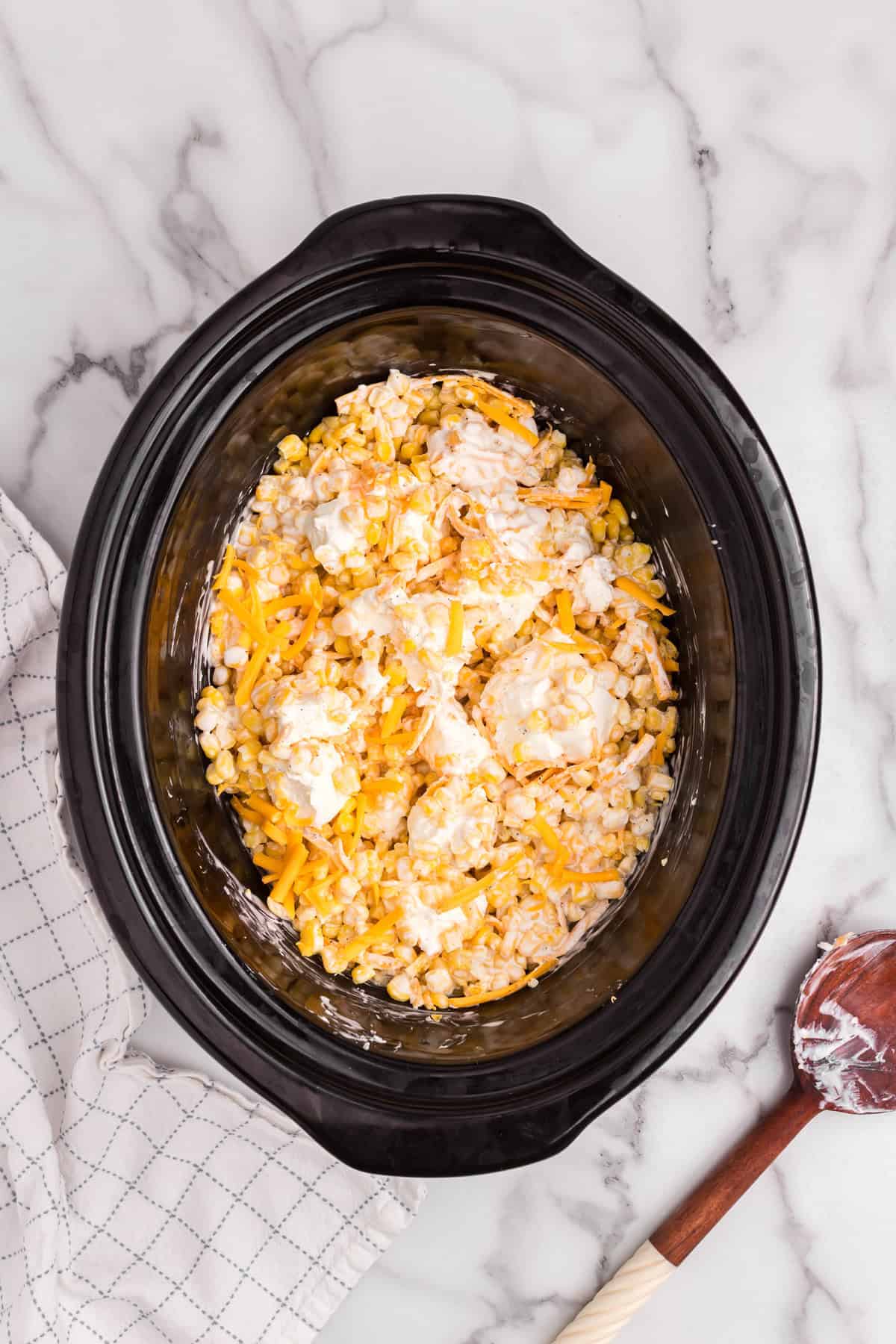 Ingredients in the slow cooker partially mixed, showing cream cheese chunks and shredded cheddar still visible among the corn and cream mixture.