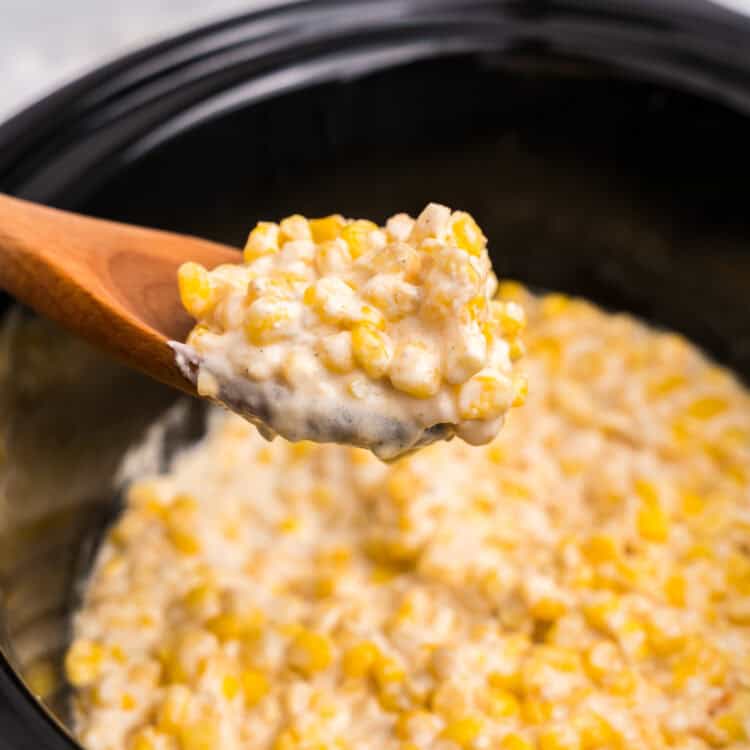 A wooden spoon lifting a portion of hot, creamy creamed corn out of the slow cooker, highlighting the thick and cheesy texture.