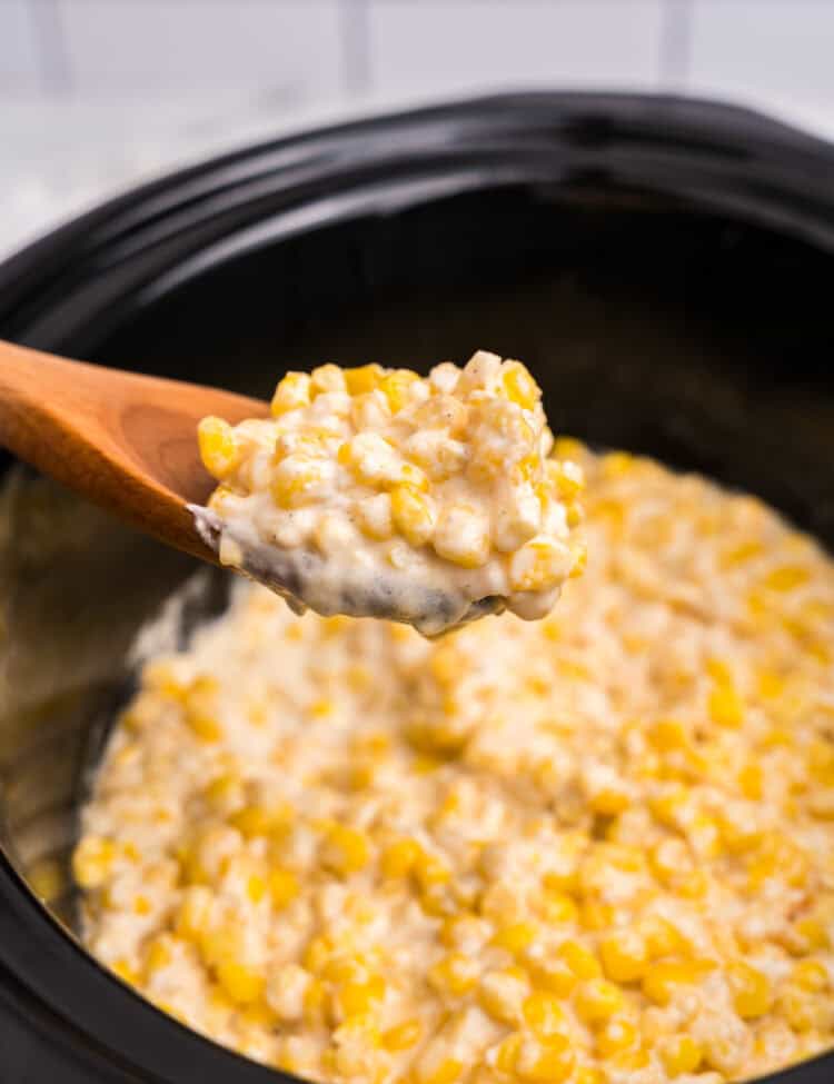A wooden spoon lifting a portion of hot, creamy creamed corn out of the slow cooker, highlighting the thick and cheesy texture.