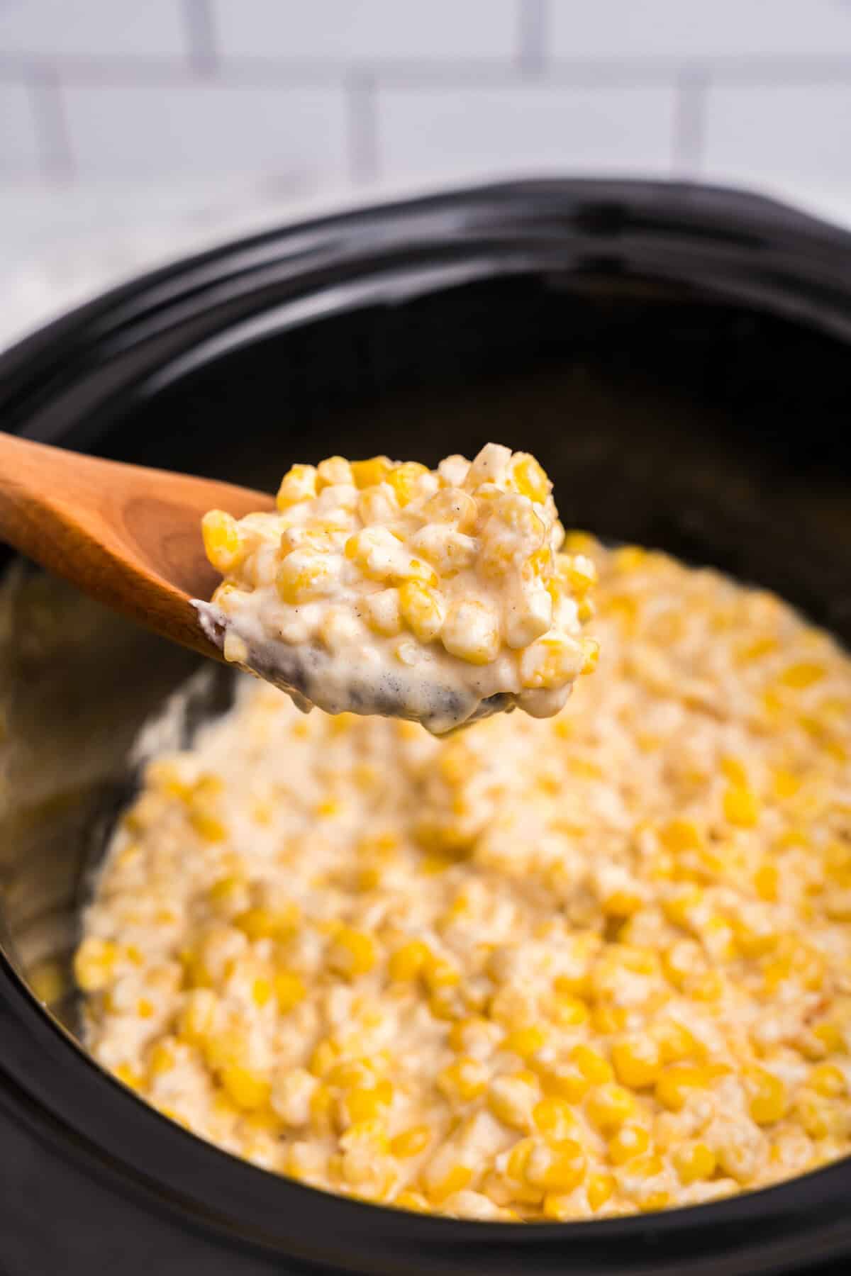 A wooden spoon lifting a portion of hot, creamy creamed corn out of the slow cooker, highlighting the thick and cheesy texture.

