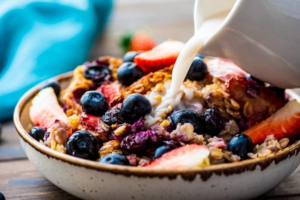 Bowl with berry baked oatmeal with a white ceramic pitcher pouring milk onto it. 