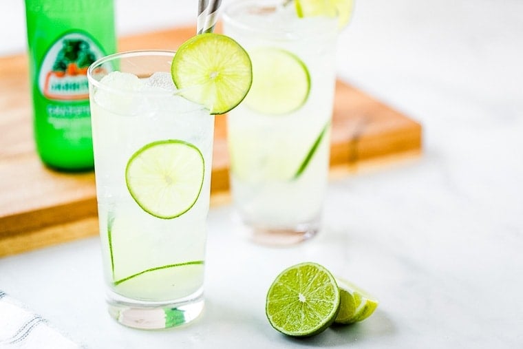 Two Paloma Cocktails with a lime on marble background. A wood cutting board in background with a bottle of green soda on it.